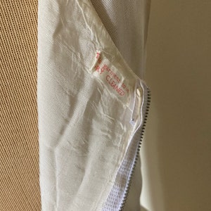 May include: A cream-colored garment featuring a silver-toned metal zipper closure. A white fabric care label is sewn near the zipper, printed with the red text: This garment to be DRY CLEANED. The fabric has a subtle, textured, woven appearance.