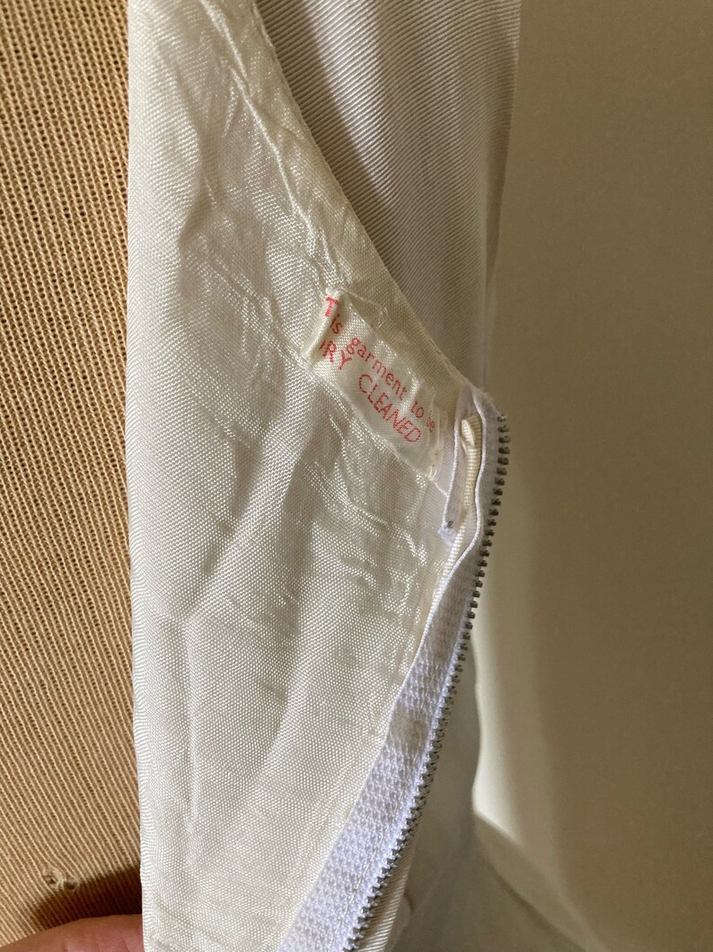 May include: A cream-colored garment featuring a silver-toned metal zipper closure. A white fabric care label is sewn near the zipper, printed with the red text: This garment to be DRY CLEANED. The fabric has a subtle, textured, woven appearance.