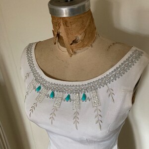 May include: A white sleeveless dress featuring a wide scoop neckline adorned with silver metallic embroidery, clear rhinestones, and dangling turquoise teardrop-shaped accents. The bodice includes delicate embroidered leaf patterns.