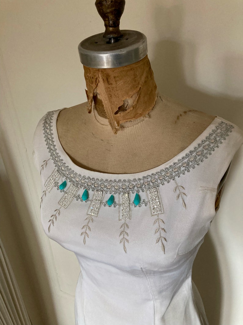 May include: A white sleeveless dress featuring a wide scoop neckline adorned with silver metallic embroidery, clear rhinestones, and dangling turquoise teardrop-shaped accents. The bodice includes delicate embroidered leaf patterns.