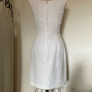 May include: A sleeveless white dress featuring a fitted bodice and a straight skirt. The garment has a high neckline and a long, centered vertical zipper closure running down the back, extending from the neckline to the lower back area.