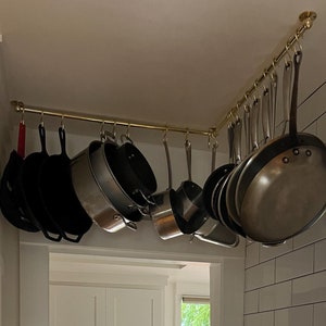 Puede incluir: Un estante de ollas de latón montado en la esquina del techo de una cocina, que contiene una variedad de ollas y sartenes, incluyendo sartenes de hierro fundido y ollas de acero inoxidable.