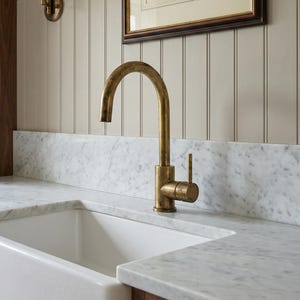 May include: A kitchen with a white ceramic sink, marble worktops, and a brass tap. A framed landscape print hangs on the wall above the sink. A wall-mounted lamp with a cream shade is also visible.