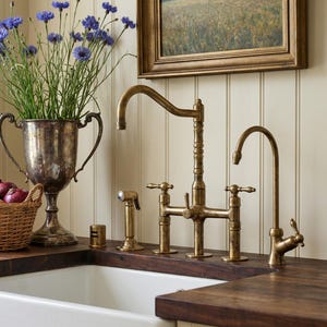 Puede incluir: Un fregadero de cocina con una pila blanca y una encimera de madera oscura. Se instala un juego de grifos de latón de estilo vintage. Un jarrón de trofeo plateado contiene flores azules. Una pintura enmarcada cuelga de la pared.
