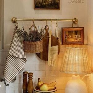 Könnte beinhalten: Eine Küchentheke mit einer weißen Fliesenrückwand, einem weißen Toaster, einer weißen Keramiklampe mit einem geflochtenen Lampenschirm, einem Holzbrett, einem geflochtenen Korb mit Lavendel, einem braunen und weißen karierten Geschirrtuch, einer Schüssel mit Obst, einem Eierkarton und zwei hölzernen Salz- und Pfefferstreuern. Die Theke ist aus Marmor und hat ein goldenes Metallgestell mit Haken, die darüber hängen.