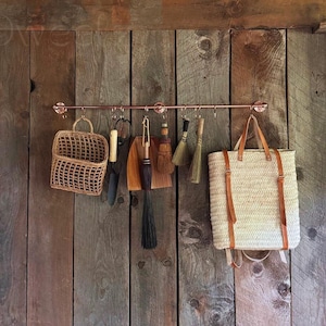 May include: A copper pipe rack mounted on a wooden wall with a variety of hanging items, including a woven basket, a straw tote bag, and several brushes.