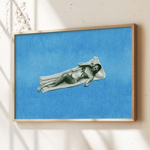 Op de afbeelding: Een ingelijste kunstdruk toont een persoon die op een zwemband ligt tegen een blauwe achtergrond. Het kunstwerk zit in een houten lijst en hangt boven een houten kast. De kamer is ingericht met planten en een neutraal gekleurde bank.