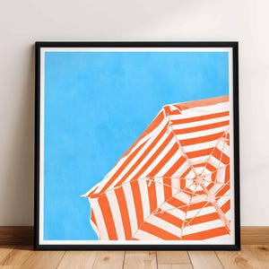 May include: Framed art print featuring a vibrant beach scene. The artwork showcases a section of a striped orange and white beach umbrella against a bright blue sky. The print is in a black frame and is displayed on a wooden floor.