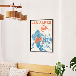 May include: A framed vintage poster with the text "LES ALPES" depicts skiers on a blue and white mountain. The poster is mounted on a white wall above a wooden bench with two decorative pillows. A wooden chandelier hangs above.