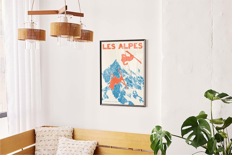 May include: A framed vintage poster with the text "LES ALPES" depicts skiers on a blue and white mountain. The poster is mounted on a white wall above a wooden bench with two decorative pillows. A wooden chandelier hangs above.