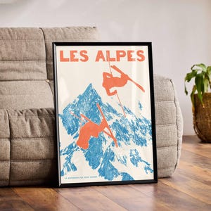 May include: Framed vintage poster with the words "LES ALPES" in orange. The poster depicts skiers in orange against a blue and white mountain backdrop. The poster is displayed on a gray sofa.