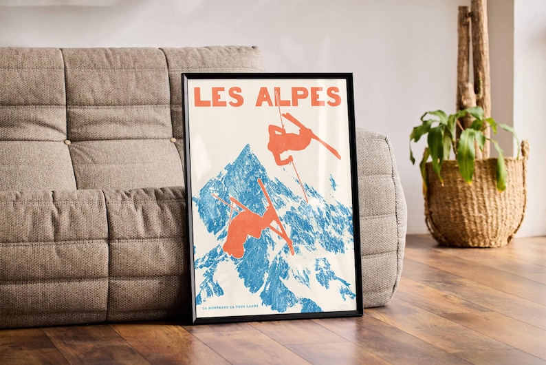 May include: Framed vintage poster with the words "LES ALPES" in orange. The poster depicts skiers in orange against a blue and white mountain backdrop. The poster is displayed on a gray sofa.