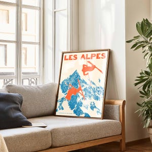 May include: A framed vintage poster with the text "LES ALPES" in red letters. The poster features a blue mountain scene with skiers in orange, set against a cream background. The poster is displayed on a light-colored sofa near a window.