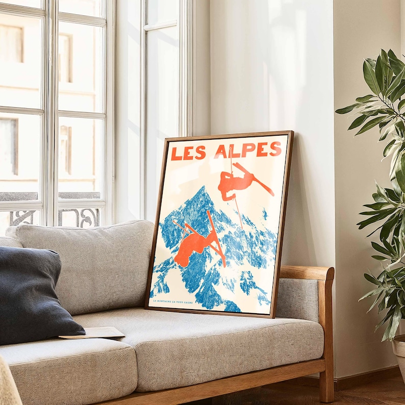 May include: A framed vintage poster with the text "LES ALPES" in red letters. The poster features a blue mountain scene with skiers in orange, set against a cream background. The poster is displayed on a light-colored sofa near a window.