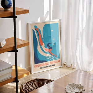 May include: A framed poster with a blue and white illustration of a person diving into water. The poster has the text "Esplora Acque Cristalline Costiera Amalfitana".