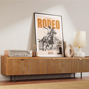 Pode incluir: Um p&ocirc;ster de estilo vintage com o texto "WELD COUNTY RODEO" em letras laranja e pretas. O p&ocirc;ster apresenta uma ilustra&ccedil;&atilde;o em preto e branco de um cowboy montando um cavalo que se encabrita. O p&ocirc;ster est&aacute; emoldurado e exposto em uma prateleira de madeira com um est&eacute;reo e uma l&acirc;mpada.