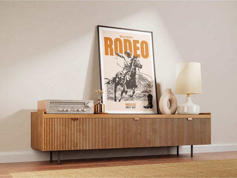 Pode incluir: Um p&ocirc;ster de estilo vintage com o texto "WELD COUNTY RODEO" em letras laranja e pretas. O p&ocirc;ster apresenta uma ilustra&ccedil;&atilde;o em preto e branco de um cowboy montando um cavalo que se encabrita. O p&ocirc;ster est&aacute; emoldurado e exposto em uma prateleira de madeira com um est&eacute;reo e uma l&acirc;mpada.