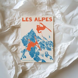 May include: A vintage-style print featuring the text "LES ALPES" in orange. The image depicts skiers in orange against a blue mountain backdrop. The print is on a cream-colored background and is surrounded by white, crumpled paper.