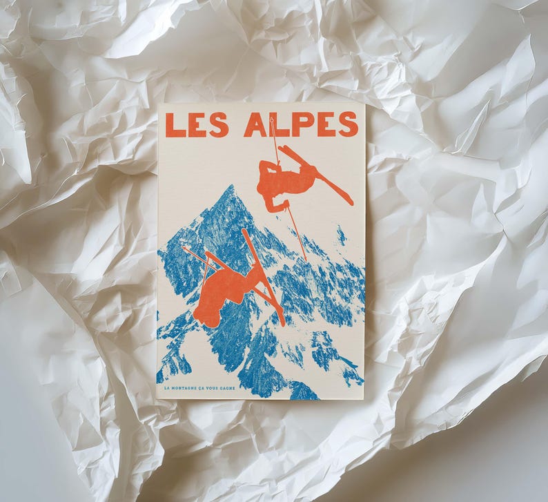 May include: A vintage-style print featuring the text "LES ALPES" in orange. The image depicts skiers in orange against a blue mountain backdrop. The print is on a cream-colored background and is surrounded by white, crumpled paper.