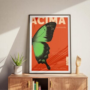 May include: Framed art print with a green and black butterfly wing on a red background. The poster has the word "ACIMA" and other Portuguese text. The artwork is displayed on a wooden cabinet, with a small plant and a wooden hand sculpture.