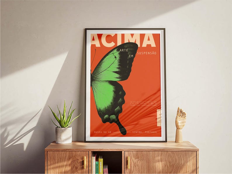May include: Framed art print with a green and black butterfly wing on a red background. The poster has the word "ACIMA" and other Portuguese text. The artwork is displayed on a wooden cabinet, with a small plant and a wooden hand sculpture.