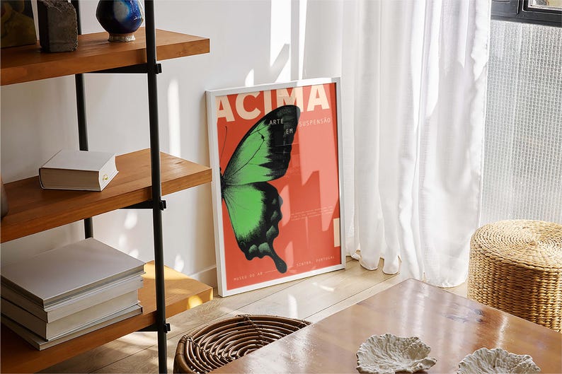 May include: A framed poster featuring a green and black butterfly wing on a red backdrop. The poster displays white text: "ACIMA ARTE EM SUSPENSO". It leans against a white wall, beside a white curtain. A wooden shelf and a woven basket are also visible.