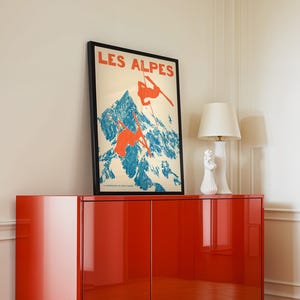 May include: A framed vintage poster with the text "LES ALPES" in red, depicting skiers on a blue and white mountain. The poster is displayed on a glossy red cabinet, with a white lamp and decorative objects.