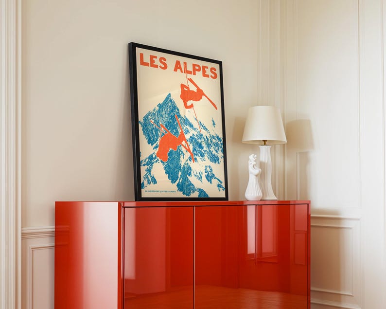 May include: A framed vintage poster with the text "LES ALPES" in red, depicting skiers on a blue and white mountain. The poster is displayed on a glossy red cabinet, with a white lamp and decorative objects.