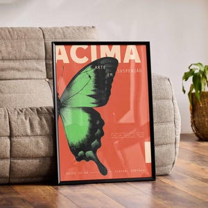 May include: Framed art print with a green and black butterfly on a red backdrop. The print features the word "ACIMA" in cream. The artwork is displayed on a grey sofa. The print is in a black frame.