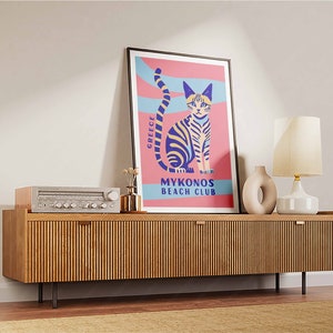 May include: A retro-style poster with a pink background and blue and white stripes. The poster features a cartoon cat with yellow eyes and a blue and yellow striped body. The text on the poster reads "Greece Mykonos Beach Club".