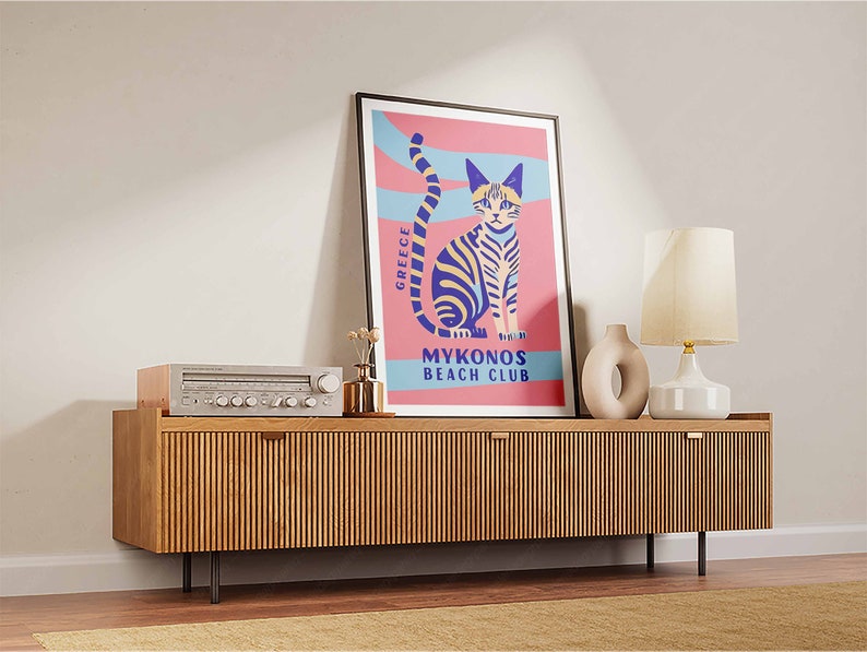 May include: A retro-style poster with a pink background and blue and white stripes. The poster features a cartoon cat with yellow eyes and a blue and yellow striped body. The text on the poster reads "Greece Mykonos Beach Club".