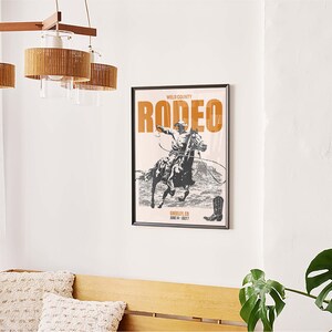 Pode incluir: Um p&ocirc;ster preto e branco com o texto "Wild County Rodeo" em letras laranja. O p&ocirc;ster apresenta um caub&oacute;i montando um cavalo. O p&ocirc;ster est&aacute; emoldurado em uma moldura preta.