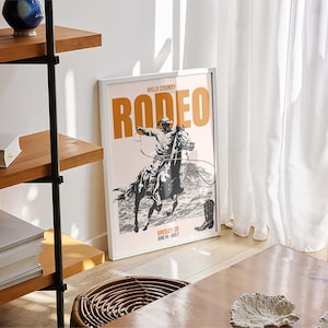 Pode incluir: Um p&ocirc;ster emoldurado branco com uma ilustra&ccedil;&atilde;o em preto e branco de um cowboy montando um cavalo. O p&ocirc;ster tem o texto "Weld County Rodeo" em laranja e o texto "Rodeo" em uma fonte laranja maior.