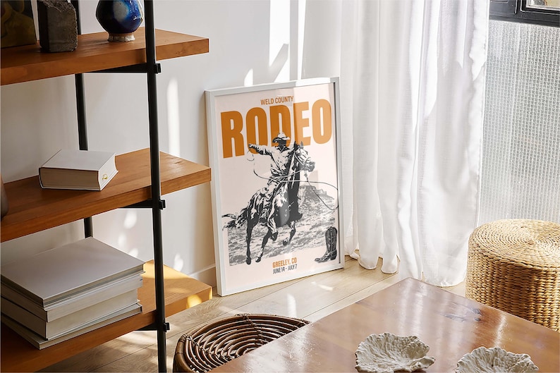 Pode incluir: Um p&ocirc;ster emoldurado branco com uma ilustra&ccedil;&atilde;o em preto e branco de um cowboy montando um cavalo. O p&ocirc;ster tem o texto "Weld County Rodeo" em laranja e o texto "Rodeo" em uma fonte laranja maior.