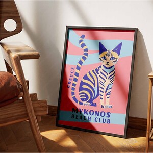 May include: A framed poster with a pink background and a stylized illustration of a cat with blue and yellow stripes. The text on the poster reads "Greece" and "Mykonos Beach Club".
