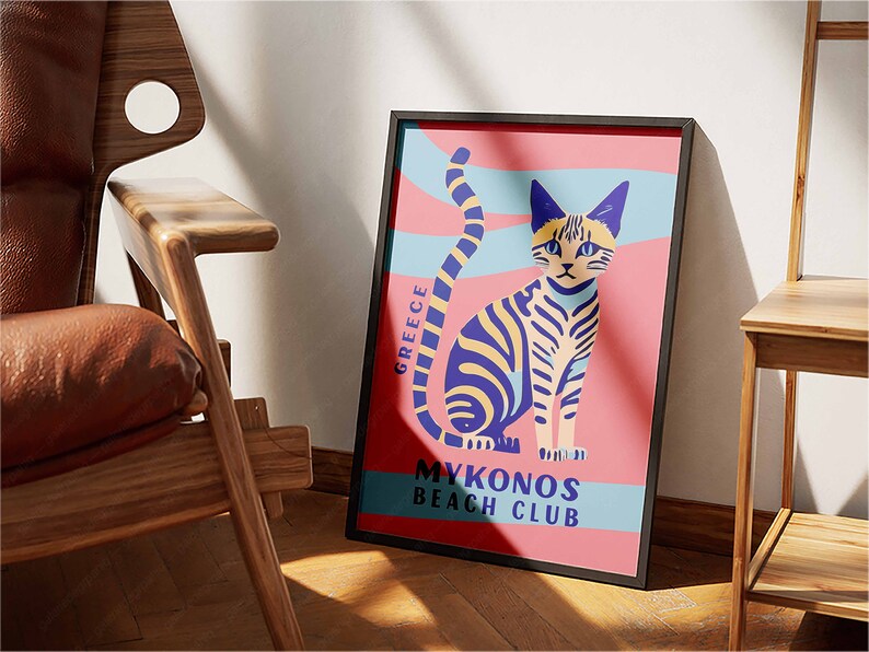 May include: A framed poster with a pink background and a stylized illustration of a cat with blue and yellow stripes. The text on the poster reads "Greece" and "Mykonos Beach Club".