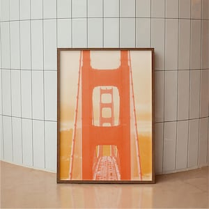 May include: A framed print of the Golden Gate Bridge in San Francisco. The bridge is depicted in orange and red hues against a yellow and beige backdrop. The print is enclosed in a dark brown wooden frame, positioned against a white tiled wall.
