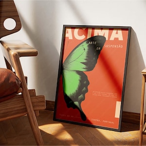 May include: Framed poster with a butterfly wing design in green and black against a red background. The poster has the word "ACIMA" at the top and other text. The poster is leaning against a white wall.