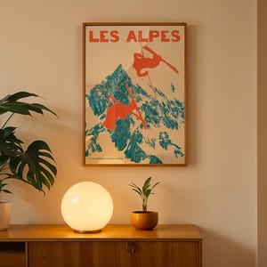 May include: A framed vintage poster with the text "LES ALPES" depicts skiers on a mountain. The poster is in a wooden frame, above a wooden cabinet. A round, glowing lamp and potted plants sit on the cabinet.