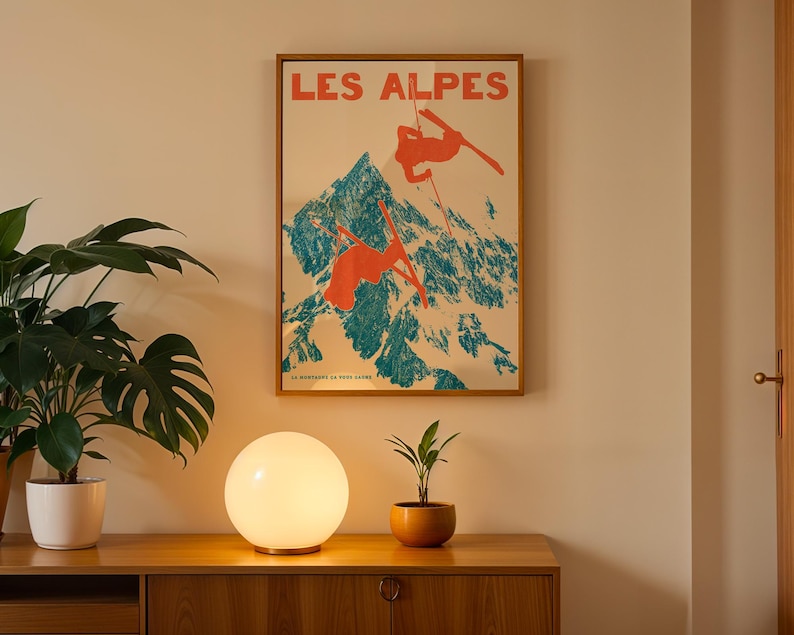 May include: A framed vintage poster with the text "LES ALPES" depicts skiers on a mountain. The poster is in a wooden frame, above a wooden cabinet. A round, glowing lamp and potted plants sit on the cabinet.