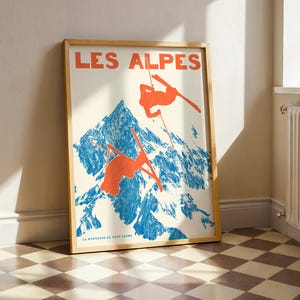 May include: Framed poster with a vintage ski resort theme. The poster features the text "LES ALPES" in orange, with illustrations of skiers in orange against a blue mountain backdrop. The poster is in a wooden frame.