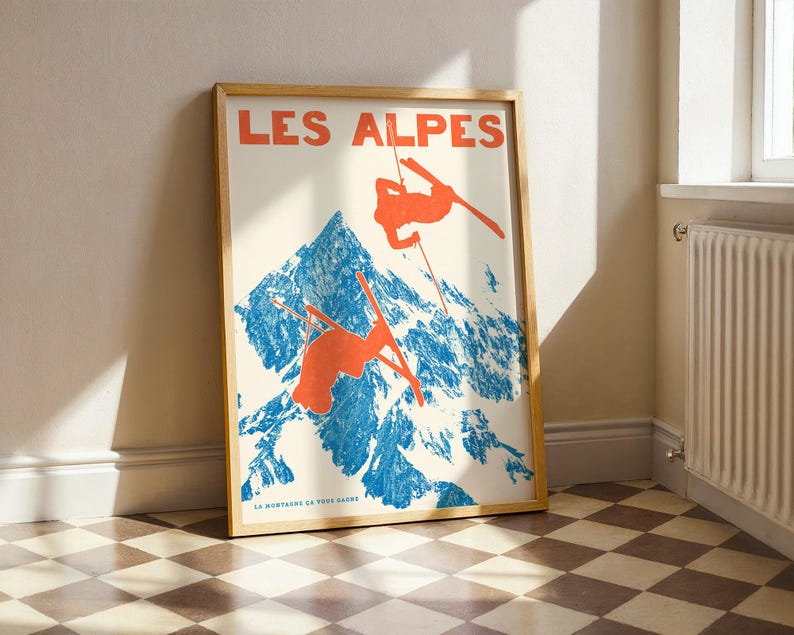 May include: Framed poster with a vintage ski resort theme. The poster features the text "LES ALPES" in orange, with illustrations of skiers in orange against a blue mountain backdrop. The poster is in a wooden frame.