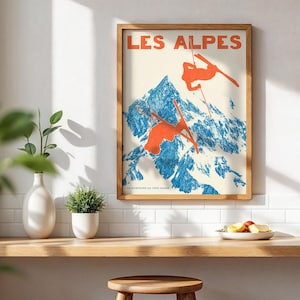 May include: A framed vintage poster with the words "LES ALPES" in red. The poster depicts skiers in orange against a blue and white mountain backdrop. The poster is in a wooden frame and is displayed on a wooden shelf.