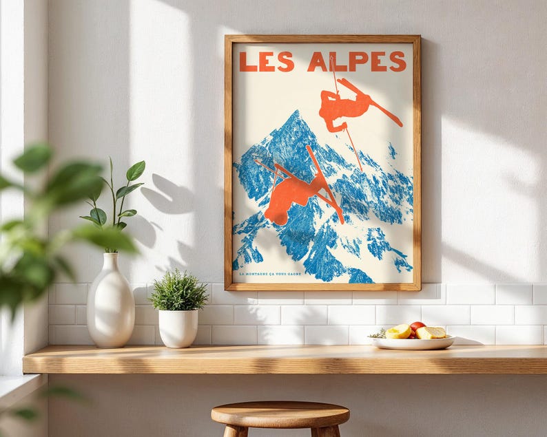 May include: A framed vintage poster with the words "LES ALPES" in red. The poster depicts skiers in orange against a blue and white mountain backdrop. The poster is in a wooden frame and is displayed on a wooden shelf.