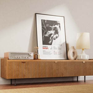 K&ouml;nnte beinhalten: Eine moderne Holz-Medienkonsole im Mid-Century-Stil mit zwei T&uuml;ren und schwarzen Metallbeinen. Die Konsole ist mit einem gerahmten Schwarzwei&szlig;-Poster eines Astronauten im Weltraum, einer wei&szlig;en Keramiklampe mit cremefarbenem Schirm und einer kleinen Vase mit Trockenblumen dekoriert.