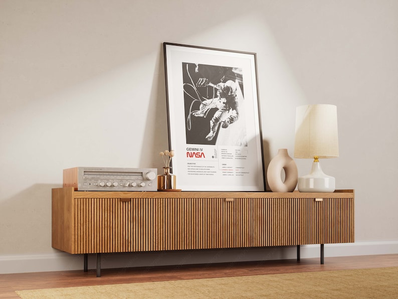 K&ouml;nnte beinhalten: Eine moderne Holz-Medienkonsole im Mid-Century-Stil mit zwei T&uuml;ren und schwarzen Metallbeinen. Die Konsole ist mit einem gerahmten Schwarzwei&szlig;-Poster eines Astronauten im Weltraum, einer wei&szlig;en Keramiklampe mit cremefarbenem Schirm und einer kleinen Vase mit Trockenblumen dekoriert.