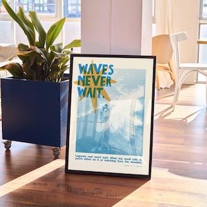 May include: Framed art print with the text "WAVES NEVER WAIT" in blue and yellow. The image depicts a surfer on a wave. The print is in a black frame and is displayed in a well-lit room with a plant and furniture.