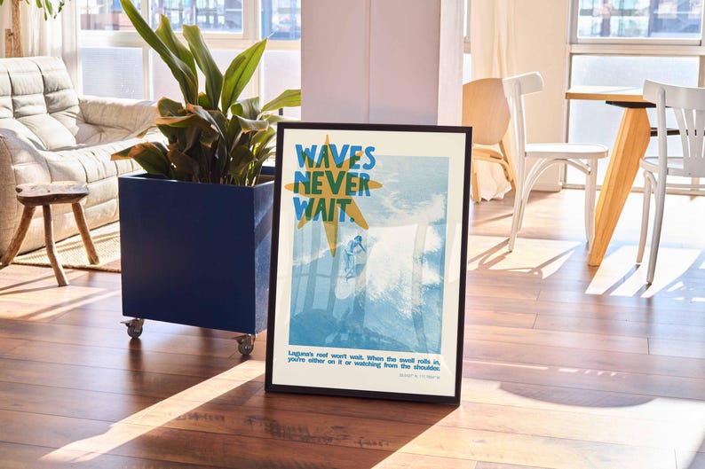 May include: Framed art print with the text "WAVES NEVER WAIT" in blue and yellow. The image depicts a surfer on a wave. The print is in a black frame and is displayed in a well-lit room with a plant and furniture.