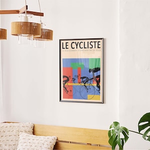 May include: A framed poster titled "LE CYCLISTE" with a colorful design of cyclists on bicycles. The poster is mounted on a white wall, above a wooden bench with decorative pillows. A modern light fixture hangs from the ceiling.