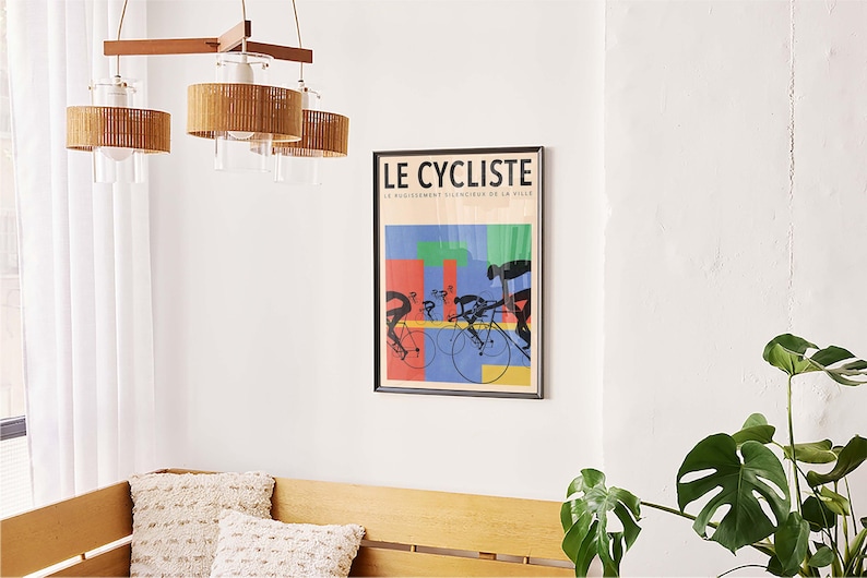 May include: A framed poster titled "LE CYCLISTE" with a colorful design of cyclists on bicycles. The poster is mounted on a white wall, above a wooden bench with decorative pillows. A modern light fixture hangs from the ceiling.